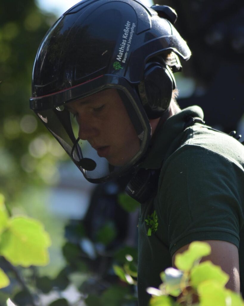 Eine Person mit Helm und Visier arbeitet konzentriert an Bäumen im Freien.